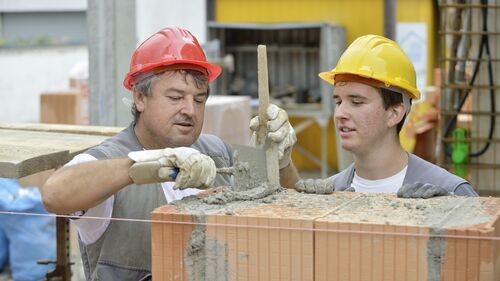 Ein Ausbilder im Maurergewerk zeigt einem Maurerlehrling, wie eine Mauer fachgerecht gesetzt wird.